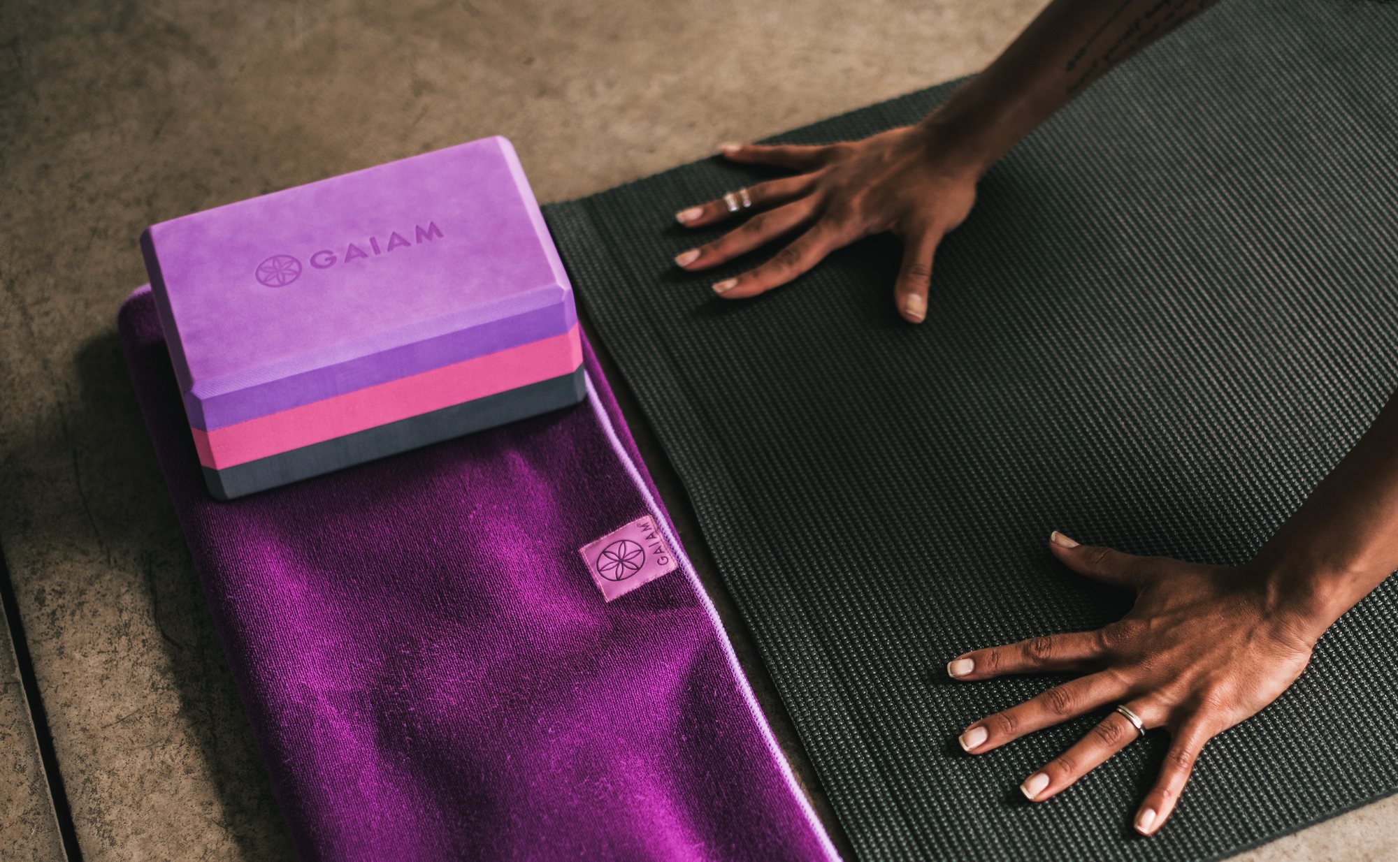 A purple Gaiam yoga block resting on a plum yoga towel beside a dark mat, with hands reaching onto the mat